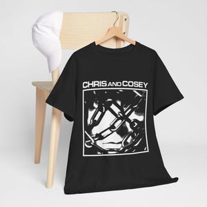 Puede incluir: Una camiseta negra de manga corta con el texto blanco CHRIS AND COSEY sobre un gráfico cuadrado de cadenas de metal entrelazadas. Una gorra de béisbol blanca lisa está colocada en el respaldo de la silla de madera detrás de la camiseta.
