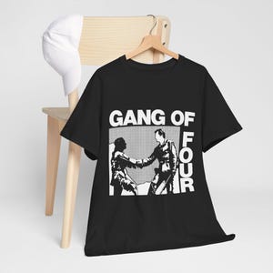 May include: A black short-sleeved t-shirt featuring a white graphic print of two figures shaking hands with the text GANG OF FOUR printed above and to the right of the image. A plain white baseball cap hangs on the back of the wooden chair.