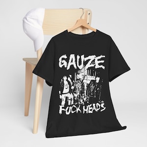 以下が含まれることがあります： 上部に「GAUZE」、下部に「FUCK HEADS」というテキストが入ったバンドの白いグラフィックプリントが施された黒の半袖Tシャツ。Tシャツは木製のハンガーに掛けられており、無地の白い野球帽が椅子の背もたれに置かれています。
