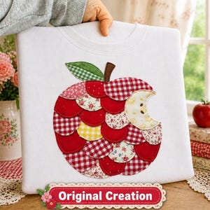 May include: A white sweatshirt featuring a large apple applique made from overlapping fabric circles in red, white, and yellow patterns, including gingham, floral, and solid prints. A green gingham leaf sits atop the apple, which has a bite mark.
