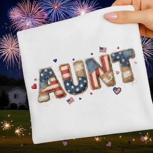 May include: A white square decorative pillow with the word "AUNT" embroidered in a patriotic design. The letters are made of fabric with American flag patterns, lace, and burlap trim. Small heart and flag accents are scattered around the letters.