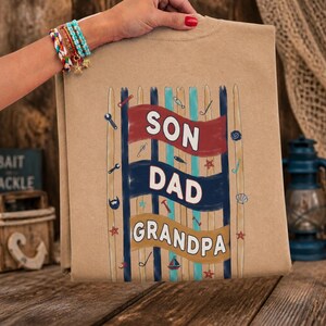 Peut inclure: Un t-shirt beige avec les mots "SON DAD GRANDPA" en blanc, rouge et beige. Le motif comprend des éléments nautiques comme des poissons, des coquillages et un voilier. Le t-shirt est tenu par une personne avec un bracelet coloré.
