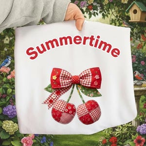 Puede incluir: Tela blanca con la palabra "Summertime" en rojo. Un lazo de cuadros rojos y blancos y dos cerezas con hojas verdes están cosidos a la tela. Las cerezas son de retazos rojos y blancos.