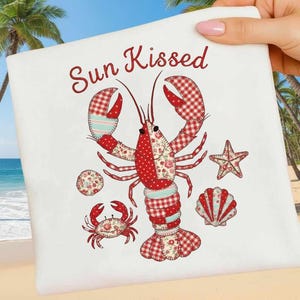 Könnte beinhalten: Weißes Strandtuch mit einem roten Hummer, einer Krabbe, einem Seestern und einem Muscheldesign. Der Hummer und andere Meeresbewohner sind aus Patchwork-Stoff gefertigt. Der Text "Sun Kissed" ist in roter Schrift geschrieben.