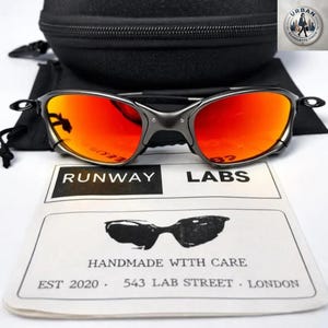 May include: Gunmetal sunglasses with vibrant orange lenses, a black case, and a drawstring bag. The sunglasses are displayed on a white surface with a card that reads "RUNWAY LABS" and "HANDMADE WITH CARE."