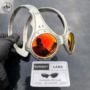 Puede incluir: Gafas de sol plateadas con lentes de color naranja. Las gafas tienen un diseño único y envolvente. Las lentes reflejan el entorno. El texto "RUNWAY LABS" y "HANDMADE WITH CARE" es visible en una etiqueta.