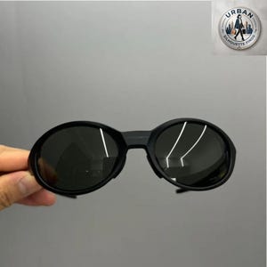 Puede incluir: Gafas de sol ovaladas negras con lentes oscuras. Las gafas tienen una montura gruesa y un puente que conecta las dos lentes. La imagen está sobre un fondo gris.
