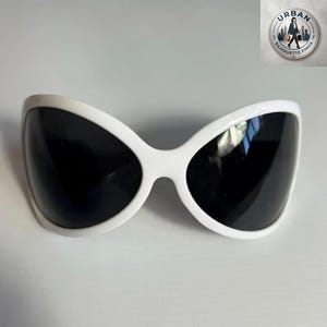 May include: White sunglasses with a bold, oversized frame and dark lenses. The sunglasses have a distinctive, curved shape, offering a modern and stylish look. The design is sleek and minimalist, perfect for adding a touch of sophistication to any outfit.