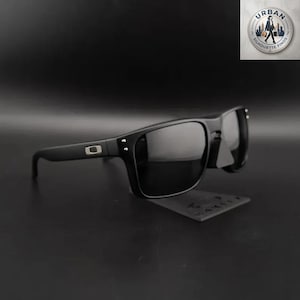 Puede incluir: Gafas de sol negras con montura cuadrada y lentes oscuras. Las gafas de sol tienen un acabado mate y un pequeño logotipo plateado en la patilla. Se muestran sobre una superficie negra.