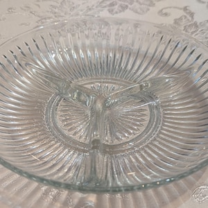 Könnte beinhalten: Klar geteilte Servierschale aus Glas mit drei Fächern. Die runde Schale hat ein geripptes Muster, das von der Mitte ausgeht. Der Mittelteil hat ein Sternmuster. Ideal zum Servieren von Snacks oder Vorspeisen.