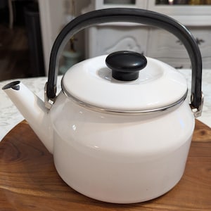 May include: A white enamel tea kettle with a black handle and a matching black knob on the lid. The kettle features a silver-toned metal rim around the top edge and a spout extending from the side. The design is simple, functional, and rounded.