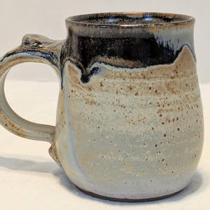 May include: A ceramic mug with a rounded body and a curved handle. The upper portion features a dark black glaze that drips down over a speckled tan and light gray base. The surface has a textured appearance with visible brown speckles throughout.