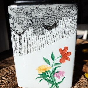 May include: A rectangular ceramic vase with a white base. The upper portion features a black and white hand-painted lace-like pattern with vertical lines. The lower portion displays a painted floral arrangement with one orange, one yellow, and one pink flower.