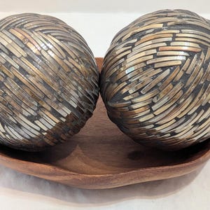 Puede incluir: Dos objetos decorativos esféricos con un patrón metálico entrelazado en tonos bronce, plata y gris oscuro, que descansan dentro de un cuenco de madera poco profundo y curvado con veta marrón natural y bordes suaves y orgánicos.