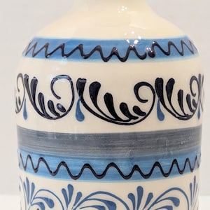 May include: A ceramic bottle with a cream-colored base featuring hand-painted horizontal bands in light blue and dark blue. The design includes decorative wavy lines and intricate swirling leaf patterns encircling the body of the vessel.