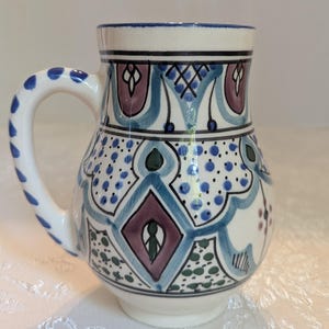 May include: A ceramic mug with a white base and handle, adorned with intricate blue, purple, and green geometric patterns. The handle is decorated with blue dots. The rim is trimmed with a blue band.
