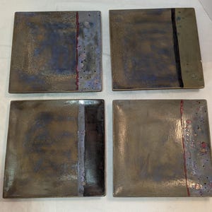 May include: Set of four square ceramic plates with a metallic bronze finish. Each plate features a vertical stripe in varying colors: red, black, and blue. The plates have a speckled design along the edges.