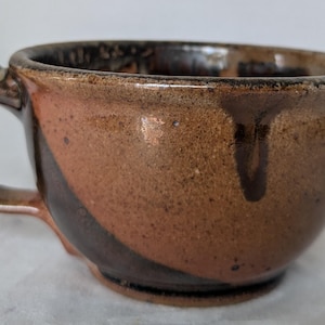 May include: A handmade ceramic mug with a rounded shape and a handle. The mug features a speckled brown and reddish-brown glaze with dark brown accents. The interior is dark brown.