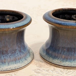 May include: Two ceramic candle holders with a flared, hourglass-shaped silhouette. The glaze features a gradient of deep navy blue at the top rim, transitioning into a lighter, mottled blue and white finish toward the wide, circular base.