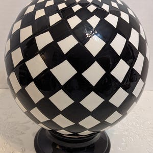 May include: A spherical ceramic object featuring a black and white checkered pattern across its surface. The sphere is mounted on a solid black circular pedestal base, creating a balanced and structured form with a glossy finish throughout.