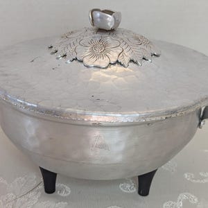 May include: Vintage aluminum serving dish with a hammered texture. The lid features a floral design and a bud-shaped handle. It has black handles and rests on three black legs. A decorative piece for serving food.