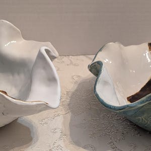 May include: Two ceramic bowls with a unique, ruffled design. One bowl is white with a dark brown accent, and the other is blue with a dark brown accent. Both bowls have a wavy, organic shape.