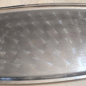 May include: A rectangular silver serving tray with decorative gold-colored handles. The tray has a slightly curved shape and ornate detailing on the corners. The surface of the tray has a textured pattern.