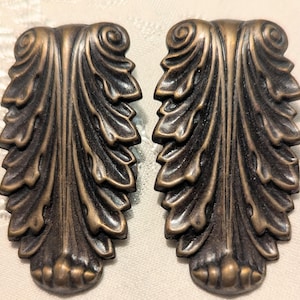 May include: Two antique bronze-toned decorative leaf-shaped cabinet pulls. Each pull features a detailed, layered leaf design with a scrolled top and a textured, rounded base. The pulls are symmetrical and have a vintage aesthetic.