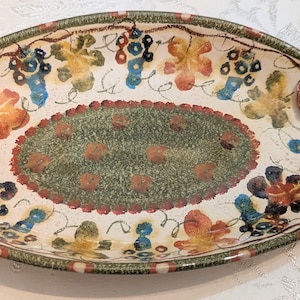 May include: An oval ceramic serving dish with two curved, textured handles. The interior features a central green oval surrounded by a painted pattern of blue grapes, yellow and orange leaves, and vines on a cream background with a green rim.