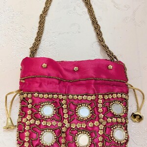 May include: A fuchsia drawstring handbag featuring a braided gold beaded handle. The front is decorated with circular mirrors, gold flower-shaped embellishments, and intricate gold beadwork. The bag closes with gold cords finished with small bell-shaped charms.