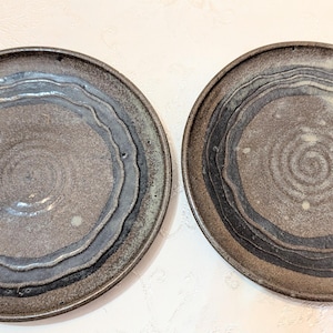 Op de afbeelding: Twee ronde keramische borden met een donkergrijze en bruine gespikkelde basis met concentrische cirkelvormige patronen in blauwe en zwarte tinten. Het oppervlak van elk bord vertoont een getextureerd uiterlijk met zichtbare ribbels.