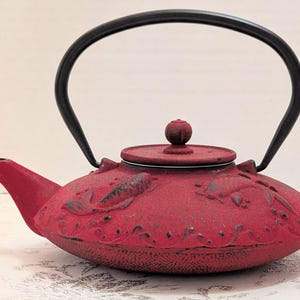 Puede incluir: Una tetera de hierro fundido roja con un asa arqueada negra, una tapa a juego con un pequeño pomo y un pico vertedor. La superficie exterior está decorada con motivos en relieve de peces koi y patrones de olas en negro.