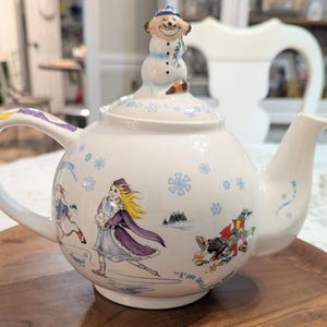 May include: A white ceramic teapot featuring illustrations of characters ice skating in a winter scene with blue snowflakes. The lid is topped with a sculpted figure of a smiling character wearing a blue hat and scarf. The text "I'm late!" is visible.