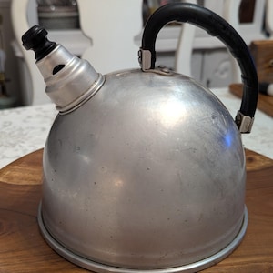 May include: A dome-shaped silver-colored metal tea kettle with a smooth finish. It features a black curved handle attached to the top and a short, tapered spout with a black stopper at the end. The kettle has a circular base and a rounded body.
