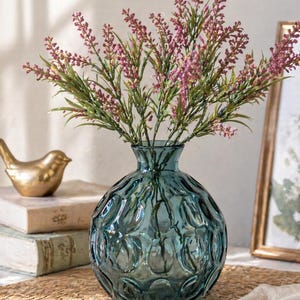 May include: A teal glass vase with a textured pattern holds a bouquet of purple and green faux flowers. A gold bird figurine and stacked books are in the background, creating a decorative home accent. The vase is approximately 20 cm tall.