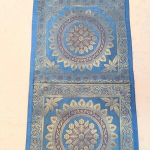 Puede incluir: Un tapiz rectangular azul que presenta dos paneles cuadrados con intrincados patrones florales de mandala en dorado y morado. La tela tiene una textura tejida, un lazo para colgar en la parte superior y un fleco decorativo en el borde inferior.