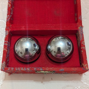 May include: A pair of shiny, silver metal balls inside a red, fabric-lined box. The box has a decorative patterned border. The balls reflect the surrounding environment. The box is open, revealing the contents.