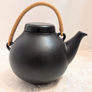 May include: A matte black ceramic teapot with a spherical body, a matching lid, and a curved spout. The teapot features a tall, arched handle wrapped in light tan woven material, which attaches to two small loops on the sides of the vessel.
