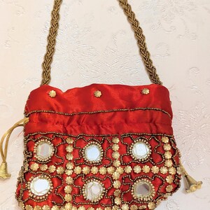 May include: A red satin drawstring pouch featuring a braided gold beaded handle. The front is decorated with circular mirrors framed by intricate gold beadwork and small gold flower-shaped metal accents. The bag closes with gold drawstrings.