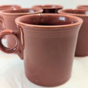 May include: A set of solid mauve-coloured ceramic mugs featuring a smooth, glossy finish. Each mug has a cylindrical body with a rounded handle on the side and subtle horizontal ring detailing near the top rim, arranged in a group.