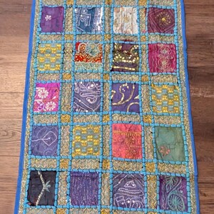 May include: A rectangular table runner featuring a grid of multicolored fabric patches in shades of red, blue, purple, green, and gold. Each square is bordered by light blue fabric strips and gold metallic trim with intricate embroidery.