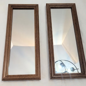 May include: A pair of rectangular wall mirrors featuring medium-toned brown wooden frames with a visible grain texture. The mirrors are positioned side-by-side, each displaying a clear reflective surface within the simple, linear frame design.