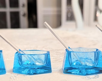 Peut inclure: Quatre petits bols hexagonaux en verre bleu translucide, chacun contenant une cuillère en verre transparent. Les bols présentent une base épaisse à facettes et un bord supérieur lisse, disposés en ligne sur une surface blanche.