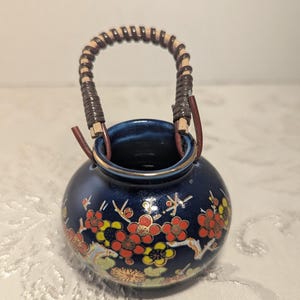Puede incluir: Un pequeño jarrón redondo de cerámica azul oscuro con un asa trenzada. El jarrón está decorado con diseños florales rojos, amarillos y dorados. El asa está hecha de un material tejido y está sujeta al jarrón con correas de cuero.
