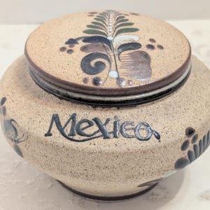 May include: A round, speckled tan ceramic jar with a matching lid. The word Mexico is hand-painted in black script on the front. The lid and body feature dark green, brown, and white floral-inspired brushwork designs with a smooth, glazed finish.