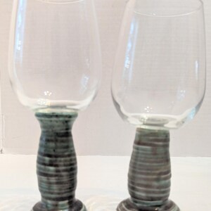 May include: Two clear glass goblets featuring thick, hand-thrown ceramic stems and bases. The stems have a textured, horizontal ribbed appearance with a mottled blue-green and grey glaze finish, supporting smooth, transparent glass bowls.