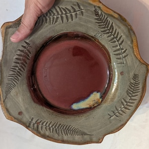 May include: Handmade ceramic plate with a deep burgundy center and a gray rim. The rim features fern leaf designs and a scalloped edge. The plate is a unique piece of pottery, perfect for serving or display.