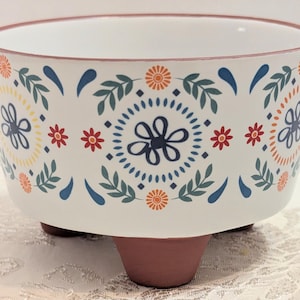 May include: A white ceramic bowl with a rounded shape and three brown feet. The exterior features a repeating pattern of dark blue flowers, orange sunbursts, and green leafy sprigs. The rim is outlined in a thin brown line, and the interior is white.