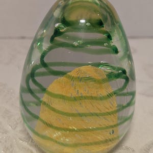 May include: A clear glass egg-shaped paperweight with a yellow and green design. The interior features a yellow, textured, egg-shaped form wrapped with green horizontal stripes and a green spiral. Small air bubbles are visible throughout the glass.