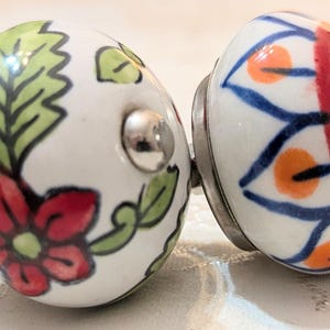 May include: Four round ceramic cabinet knobs with white bases and distinct hand-painted designs. The patterns include red and blue abstract shapes, a red flower with green leaves, a blue and orange geometric motif, and dark green leafy branches.